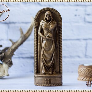 May include: A wooden carving of Hel, the Norse goddess of the underworld, depicted in a hooded robe and holding a skull. The carving is on a wooden base with the inscription "NM".