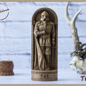 May include: A wooden carving of the Norse god Tyr, standing in a carved wooden frame. The carving is detailed and shows Tyr in full armor, holding a sword. The base of the carving is inscribed with the runes for Tyr.