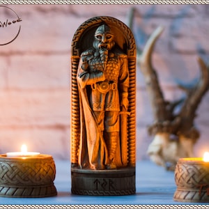 May include: A wooden carving of a Viking warrior with a beard and helmet, standing in a carved wooden frame. The carving is on a wooden base with the rune "Tyr" carved into it. The carving is lit by two candles in wooden candle holders.