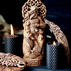 May include: A wooden carved statue of a bearded figure holding a spear, with intricate knotwork and animal carvings. Two black honeycomb candles with lit flames flank the statue, alongside other wooden decorative pieces.