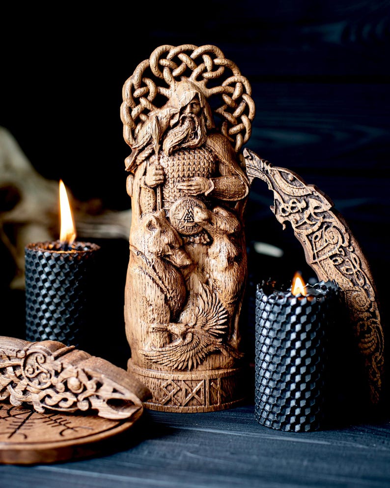 May include: A wooden carved statue of a bearded figure holding a spear, with intricate knotwork and animal carvings. Two black honeycomb candles with lit flames flank the statue, alongside other wooden decorative pieces.