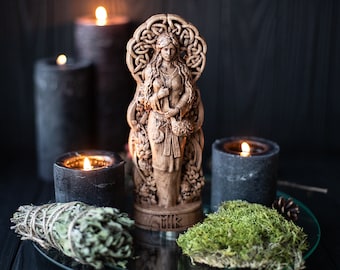 May include: A wooden statue of a female figure with long hair, standing in front of a Celtic knot design. The statue is surrounded by black candles and a bundle of dried herbs.