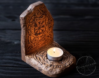 Puede incluir: Un portavelas de madera con un diseño tallado de una brújula y otros símbolos. Una pequeña vela blanca está encendida dentro del portavelas.