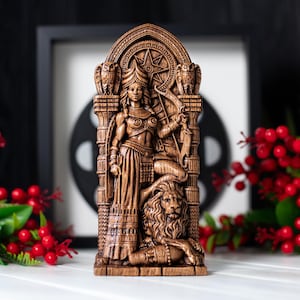 May include: A detailed wooden carving of a female figure holding a bow and arrow, flanked by owls. The figure is standing above a lion. The carving is brown and set against a blurred background.