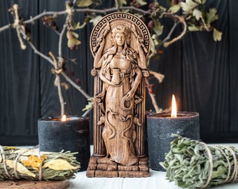 May include: Wooden statue of a goddess with multiple arms holding a cup, a snake, and a staff. The statue is carved with intricate details and stands on a base. Two black candles are on either side of the statue.