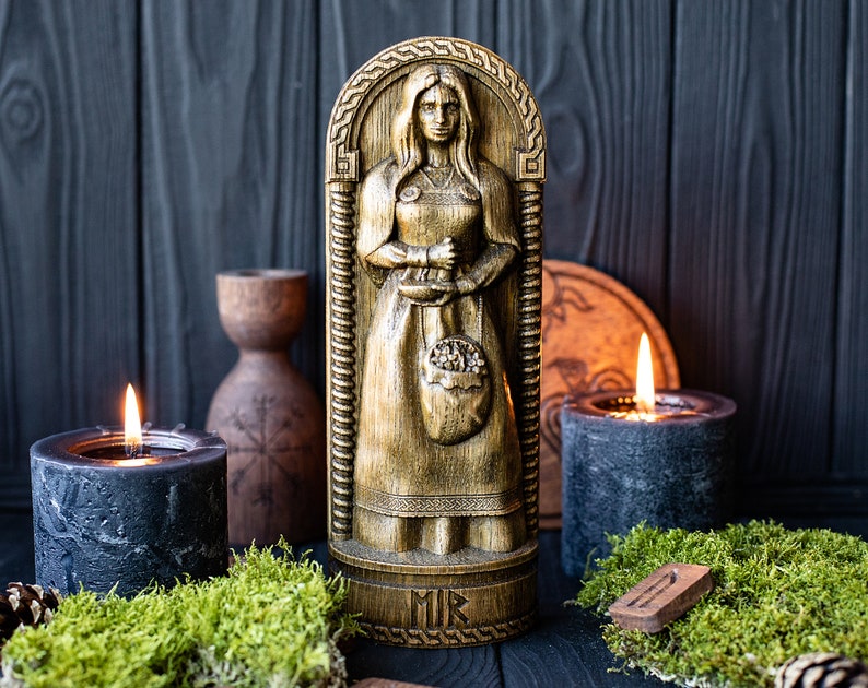 May include: A wooden carving of a woman holding a bag of grain. The carving is painted gold and has a dark wood base with the word "MIR" inscribed on it. The carving is surrounded by candles and moss.