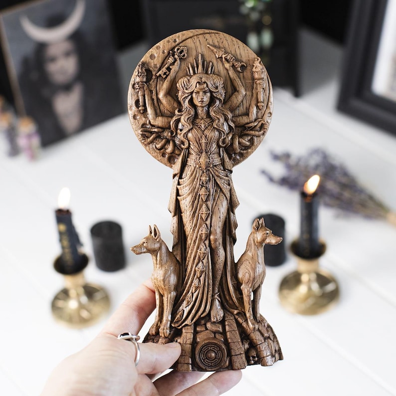 Op de afbeelding: Een gedetailleerd houten beeld van een godin met meerdere armen, een kroon en vloeiende gewaden. Twee hondfiguren flankeren de godin. Het beeld is uit hout gesneden en staat op een getextureerde basis. De achtergrond bevat kaarsen en een ingelijste foto.