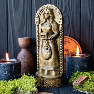 May include: A wooden carving of a woman holding a basket of fruit. The carving is painted in gold and brown tones and is set against a dark wood background. There are two black candles and moss in the foreground.