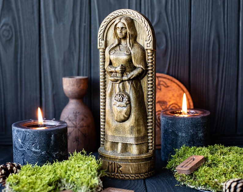 May include: A wooden carving of a woman holding a basket of fruit. The carving is painted in gold and brown tones and is set against a dark wood background. There are two black candles and moss in the foreground.