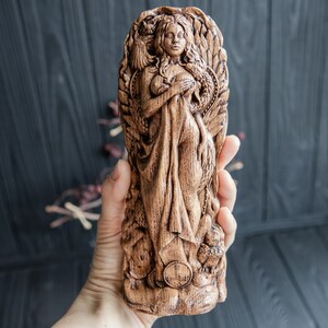 May include: A wooden carving of a woman with long hair and a flowing robe. The carving is detailed and intricate, with a focus on the woman's face and hands. The carving is set against a dark wooden background.