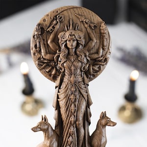Op de afbeelding: Een gedetailleerd houten beeld van een godin met meerdere armen, een kroon en vloeiend haar. De figuur staat tegen een cirkelvormige achtergrond met gesneden details. Twee hondfiguren staan aan de voet van het beeld.