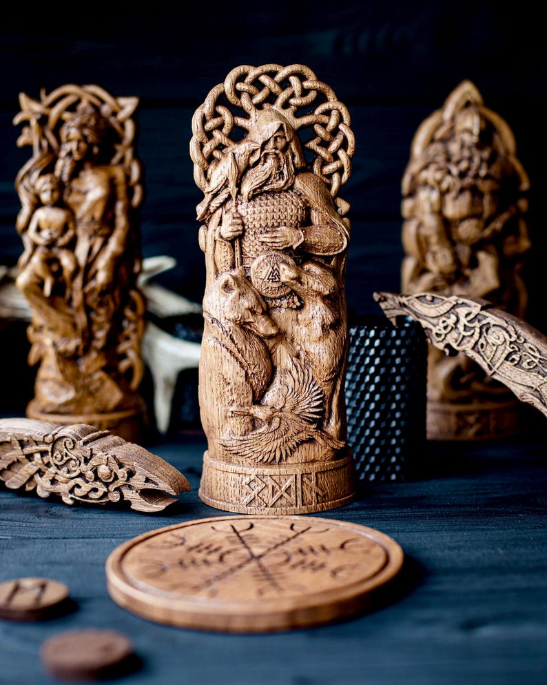 May include: Wooden carvings of Norse gods and symbols. The central figure is a detailed sculpture of a bearded man with bears, a shield, and a bird. Other carvings include a woman with a child, a wolf, and a round wooden piece with a symbol.
