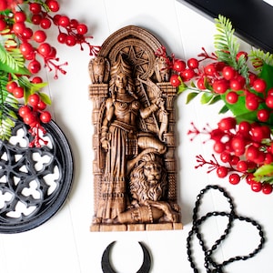 May include: A detailed, brown, carved wooden wall hanging depicting a woman with a bow and arrow, standing on a lion. The artwork features an archway design with owls and a star. Decorative red berries and black accents surround the artwork.