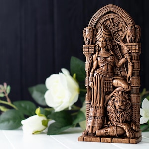 May include: A detailed wooden statue of a woman holding a bow, with a lion at her feet. The statue is brown with intricate carvings, including a star and owl details. The background is a dark wooden surface with white flowers.