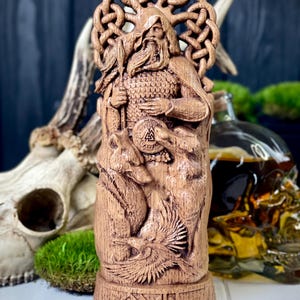 May include: A detailed wooden carving of a bearded figure with a circular halo, holding a staff and shield. The carving features intricate details of animals and symbols, with a base adorned with geometric patterns. The statue is a light brown color.