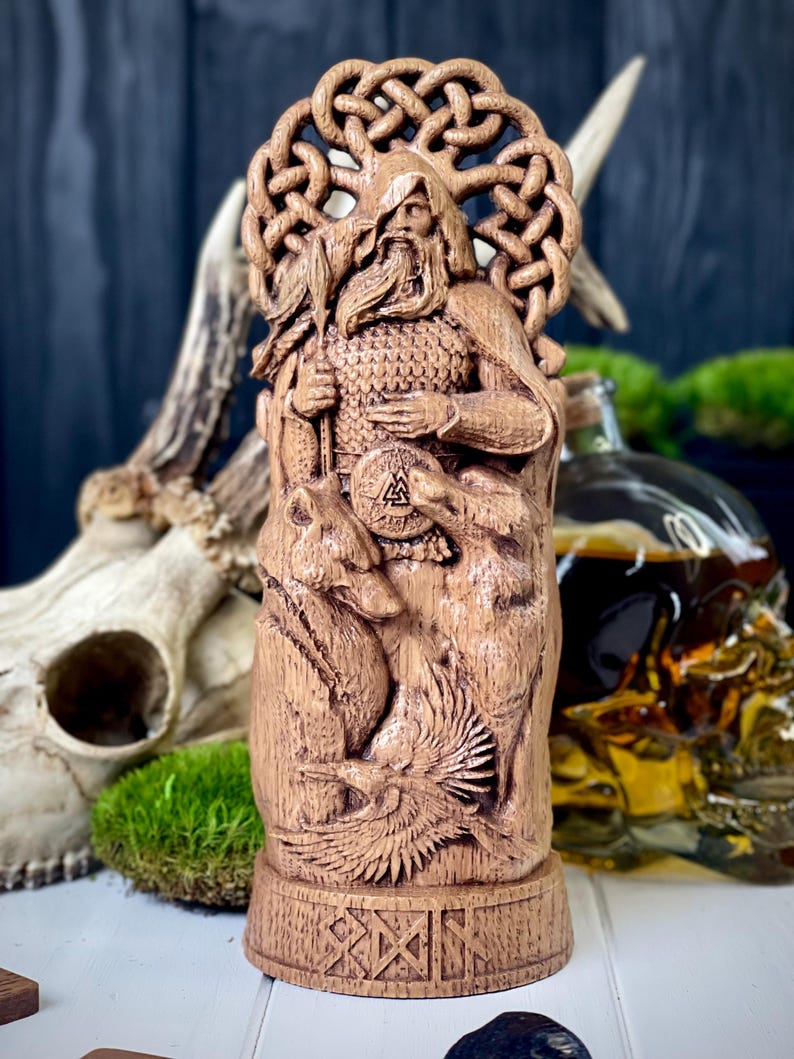 May include: A detailed wooden carving of a bearded figure with a circular halo, holding a staff and shield. The carving features intricate details of animals and symbols, with a base adorned with geometric patterns. The statue is a light brown color.