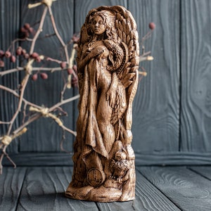 May include: A wooden sculpture of a woman with wings, holding a bird in her arms. The sculpture is carved in a detailed style and has a dark brown finish.