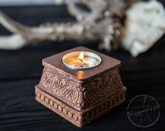 May include: A wooden candle holder with intricate carvings in a Celtic knot design. The holder is square and has a small, round indentation in the center to hold a tea light candle. The candle is lit and glowing.