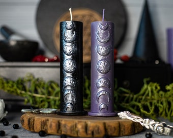 May include: Two pillar candles with a moon phase design. One candle is black with silver moon phases and the other is purple with silver moon phases.