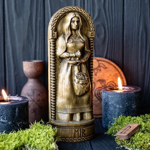 May include: A wooden carving of a woman holding a basket of flowers. The carving is painted gold and has a dark wood background. The carving is labeled "MIR" at the bottom.