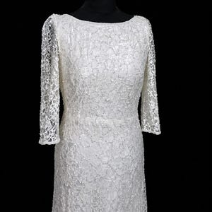Puede incluir: Un vestido de novia blanco, largo, con cuello barco y mangas largas. El vestido est&aacute; hecho de una delicada tela de encaje, con detalles intrincados. Las mangas son ajustadas y terminan en las mu&ntilde;ecas. El vestido se exhibe en un maniqu&iacute; negro.