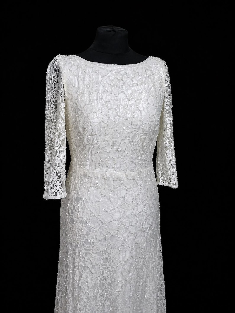 Puede incluir: Un vestido de novia blanco, largo, con cuello barco y mangas largas. El vestido est&aacute; hecho de una delicada tela de encaje, con detalles intrincados. Las mangas son ajustadas y terminan en las mu&ntilde;ecas. El vestido se exhibe en un maniqu&iacute; negro.