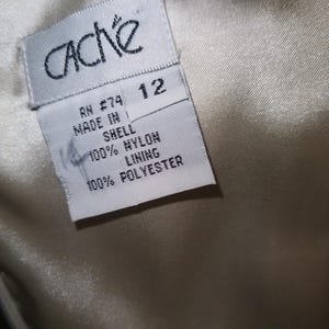 May include: A white clothing tag with black text attached to a cream-colored fabric. The tag reads CACHE, size 12, RN 74, MADE IN, SHELL 100% NYLON, and LINING 100% POLYESTER. The fabric has a smooth, satin-like texture and a subtle sheen.