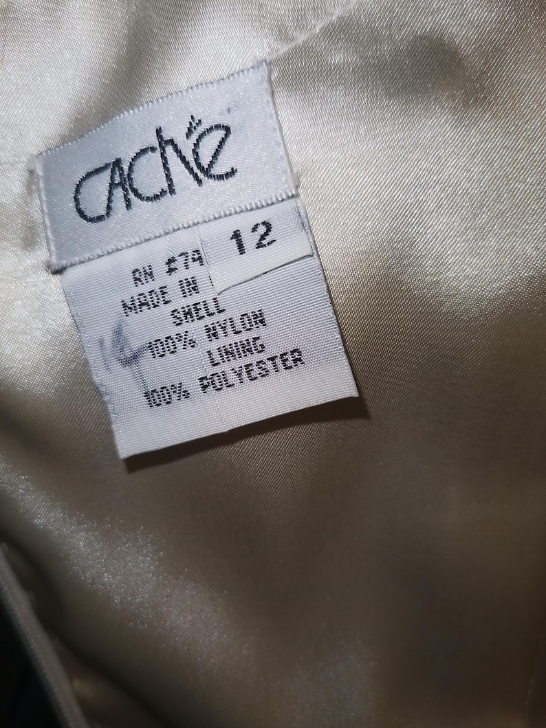 May include: A white clothing tag with black text attached to a cream-colored fabric. The tag reads CACHE, size 12, RN 74, MADE IN, SHELL 100% NYLON, and LINING 100% POLYESTER. The fabric has a smooth, satin-like texture and a subtle sheen.
