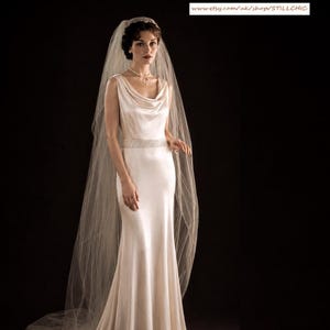 May include: A floor-length ivory satin gown featuring a cowl neckline, a sleeveless bodice, and a beaded waist detail. The garment is paired with a long, sheer, floor-sweeping tulle veil that drapes over the shoulders and down the back.
