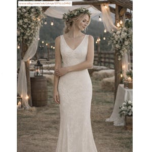 May include: A sleeveless ivory wedding dress with a V-neck and a fitted silhouette. The dress features an all-over floral lace pattern. The bride is wearing a floral crown. The background includes a wooden archway, string lights, and floral arrangements.