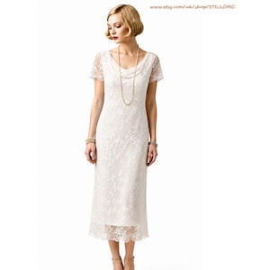 May include: A cream-colored midi-length dress featuring short sleeves, a cowl neckline, and an intricate floral lace overlay. The garment is styled with a long beaded necklace and a bracelet. The text www.etsy.com/uk/shop/STILLCHIC is visible.