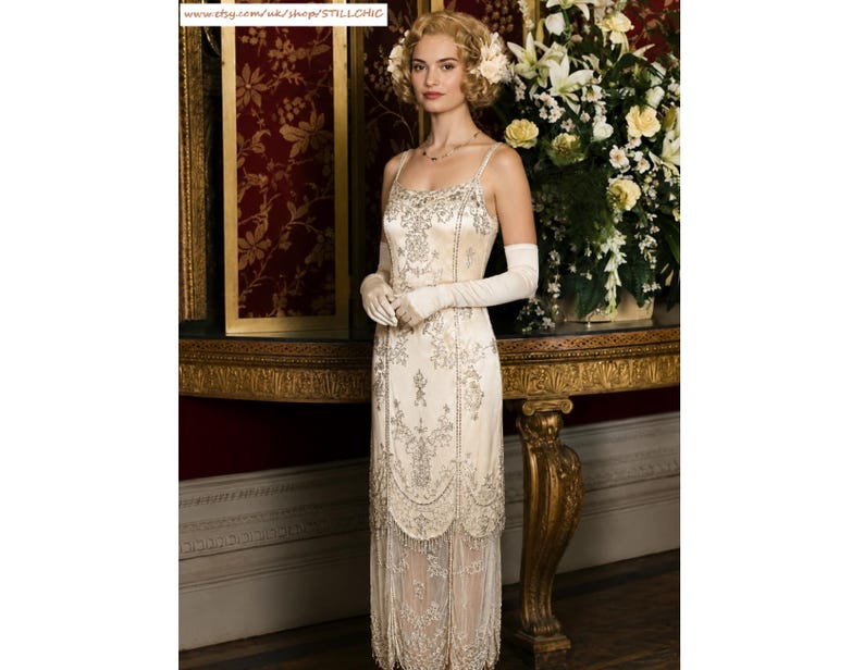 May include: A cream-colored sleeveless dress featuring intricate silver beadwork and embroidery throughout the bodice and skirt, finished with a scalloped hemline and a long, layered fringe detail that extends to the lower calf area.