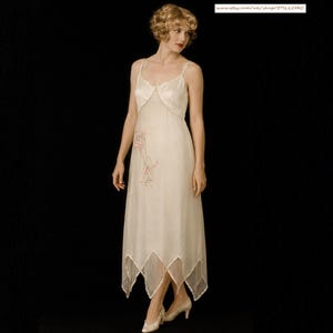 May include: A sleeveless, floor-length cream-colored dress with thin spaghetti straps and a V-neckline. The dress features a delicate pink floral embroidery on the bodice and a handkerchief hemline. The fabric appears lightweight and sheer.