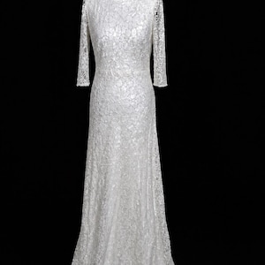 Puede incluir: Vestido largo de encaje blanco con mangas largas y cuello barco. El vestido se exhibe en un maniqu&iacute; negro sobre un fondo negro. El patr&oacute;n de encaje es intrincado y cubre todo el vestido.