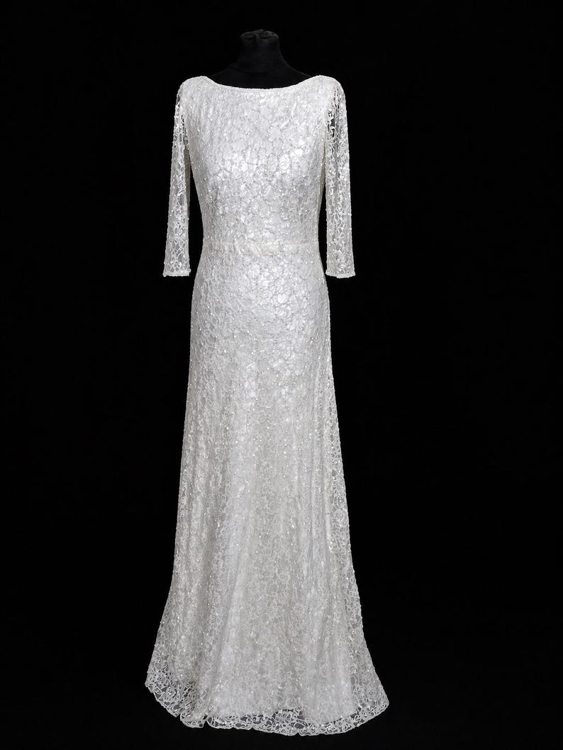 Puede incluir: Vestido largo de encaje blanco con mangas largas y cuello barco. El vestido se exhibe en un maniqu&iacute; negro sobre un fondo negro. El patr&oacute;n de encaje es intrincado y cubre todo el vestido.