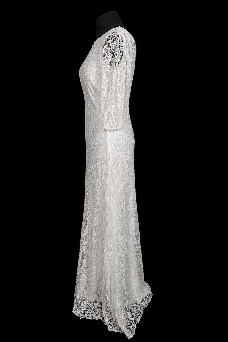 Puede incluir: Un vestido blanco largo con una delicada capa de encaje. El vestido presenta mangas largas y un escote en V sutil. Los detalles de encaje se extienden a las mangas y al dobladillo, creando un look rom&aacute;ntico y elegante. El vestido se muestra en un maniqu&iacute; negro.