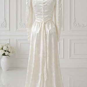 K&ouml;nnte beinhalten: Ein elfenbeinfarbenes Vintage-Brautkleid mit langen &Auml;rmeln und hohem Ausschnitt. Das Kleid hat ein eng anliegendes Oberteil mit Kn&ouml;pfen am R&uuml;cken und einen flie&szlig;enden Rock. Ein passender G&uuml;rtel wird in der Taille gebunden. Das Kleid wird auf einer Schaufensterpuppe pr&auml;sentiert.