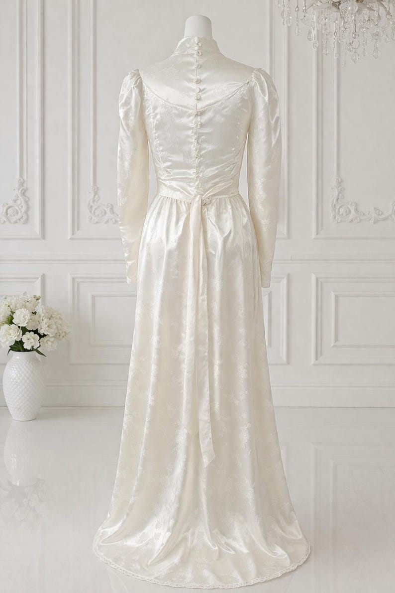 K&ouml;nnte beinhalten: Ein elfenbeinfarbenes Vintage-Brautkleid mit langen &Auml;rmeln und hohem Ausschnitt. Das Kleid hat ein eng anliegendes Oberteil mit Kn&ouml;pfen am R&uuml;cken und einen flie&szlig;enden Rock. Ein passender G&uuml;rtel wird in der Taille gebunden. Das Kleid wird auf einer Schaufensterpuppe pr&auml;sentiert.