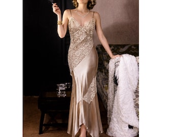 May include: A floor-length champagne-colored satin slip dress featuring asymmetrical panels of intricate white lace. The garment has thin spaghetti straps, a V-neckline, and a flared hemline that creates a fluid silhouette with a subtle sheen.