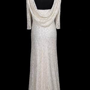 Puede incluir: Vestido largo de color marfil con cuello drapeado y mangas tres cuartos. El vestido est&aacute; cubierto con un delicado patr&oacute;n texturizado y presenta una sutil cola. El vestido se exhibe en un maniqu&iacute; negro sobre un fondo negro.