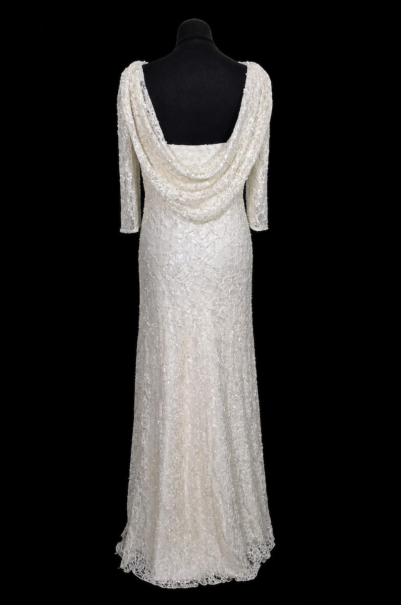 Puede incluir: Vestido largo de color marfil con cuello drapeado y mangas tres cuartos. El vestido est&aacute; cubierto con un delicado patr&oacute;n texturizado y presenta una sutil cola. El vestido se exhibe en un maniqu&iacute; negro sobre un fondo negro.
