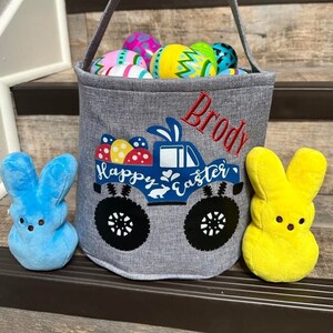 May include: A gray Easter basket with a monster truck design and the text "Happy Easter" and "Brody". The basket is filled with colorful Easter eggs and is sitting on a set of stairs. There are two blue and yellow plush bunny toys on either side of the basket.