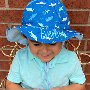 May include: A blue and white sun hat with a shark pattern and the name "Everett" embroidered on the front. The hat has a wide brim and a chin strap.