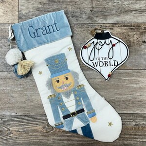 May include: A white Christmas stocking with a blue velvet cuff embroidered with the name "Grant". The stocking features a nutcracker design with gold accents and a white pom-pom. A white wooden ornament with the text "Joy to the World" is also pictured.