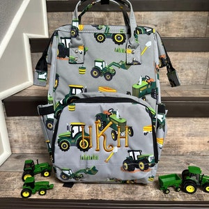 May include: Gray backpack with a green tractor print. The backpack has a large front pocket and a zipper closure. The backpack is monogrammed with the initials "HKH".