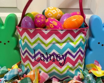May include: A pink and green chevron Easter basket with the name "Lainey" embroidered on the front. The basket is filled with colorful plastic Easter eggs.