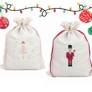 May include: Two white fabric gift bags with red drawstrings. One bag features an embroidered ballerina, the other a nutcracker. Both bags are decorated with gold glitter and are surrounded by colorful Christmas ornaments.