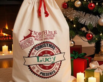 May include: A cream-colored fabric gift sack with a red drawstring closure. The front features a red and black graphic with the text North Pole Post Office Special Delivery, Christmas Presents, and the name Lucy printed in a central banner.