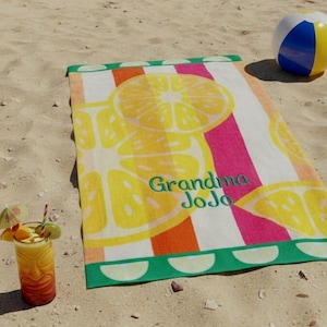 May include: A rectangular beach towel featuring a vibrant pattern of yellow lemon slices over vertical stripes of pink, orange, and white. The text "Grandma JoJo" is embroidered in green script in the center, with a green border featuring white semi-circles.