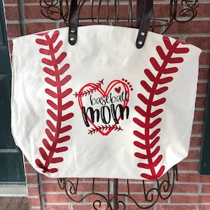 May include: A white canvas tote bag with red baseball stitching and a red heart with the text "baseball mom" inside. The bag has brown leather handles.